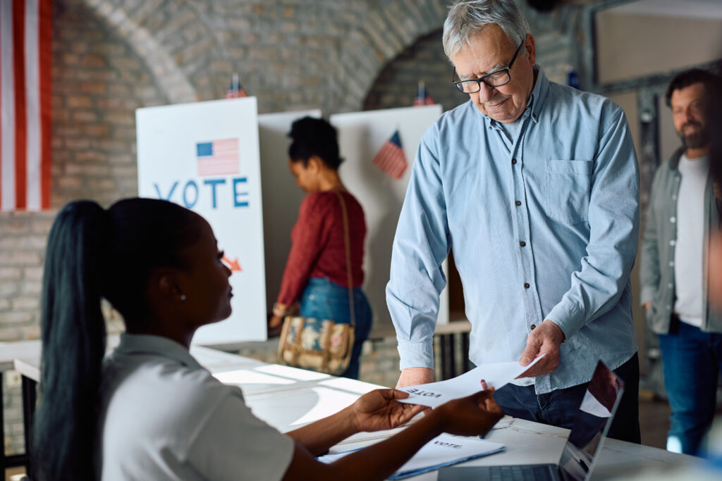 Senior US citizen receiving ballot from polling place volunteer during election voting.