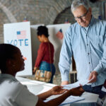 Senior US citizen receiving ballot from polling place volunteer during election voting.