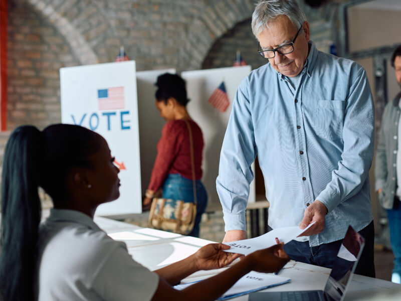 Senior US citizen receiving ballot from polling place volunteer during election voting.